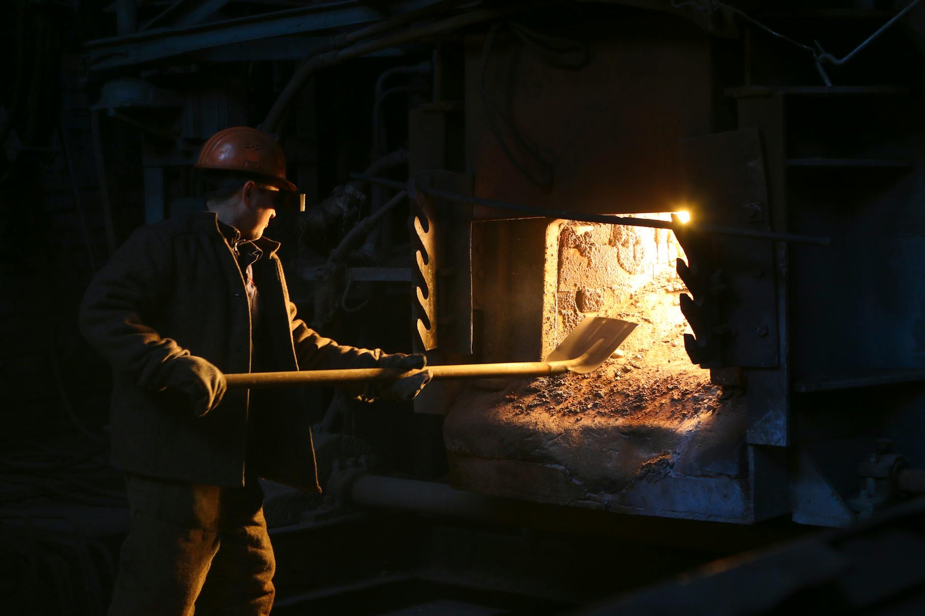 A man in a hard hat works with a furnace using a shovel, showcasing industrial labor.