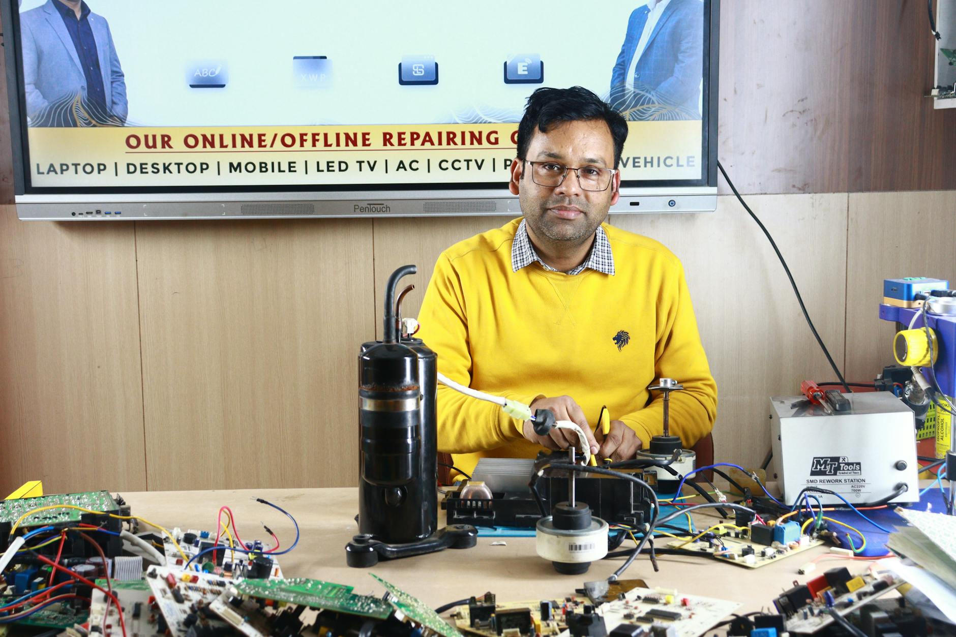 Man engages in air conditioner repair at New Delhi training center, surrounded by electronic components.