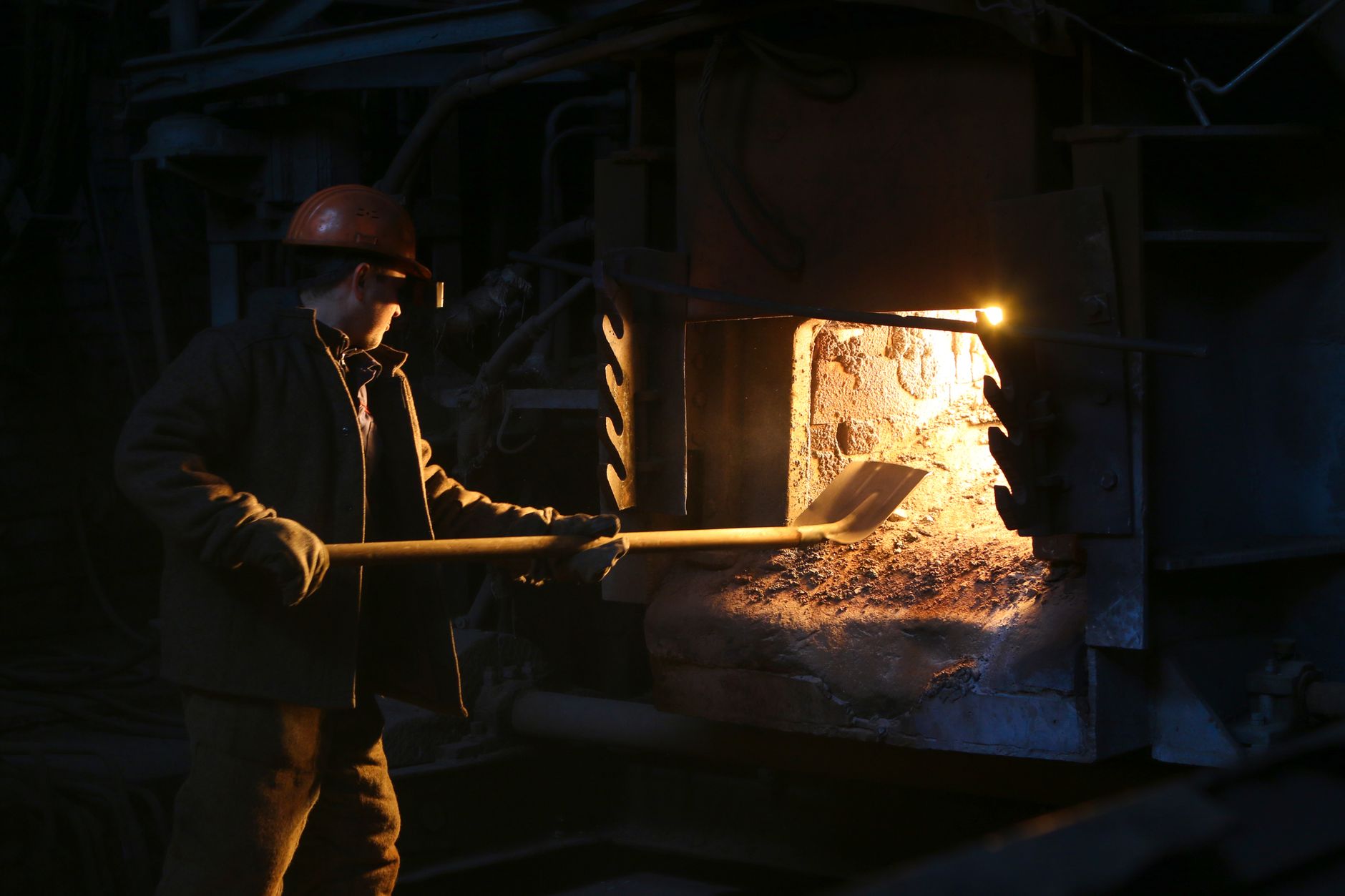 A man in a hard hat works with a furnace using a shovel, showcasing industrial labor.